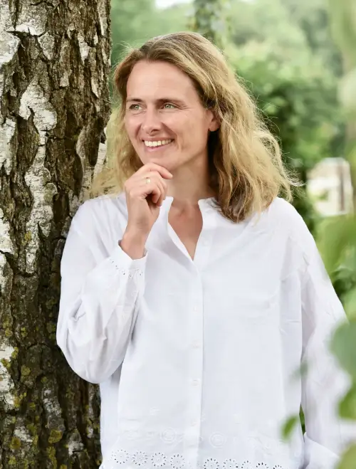 Coachin lehnt entspannt an einem Baum und blickt nachdenklich zur Seite, natürliche Umgebung im Hintergrund