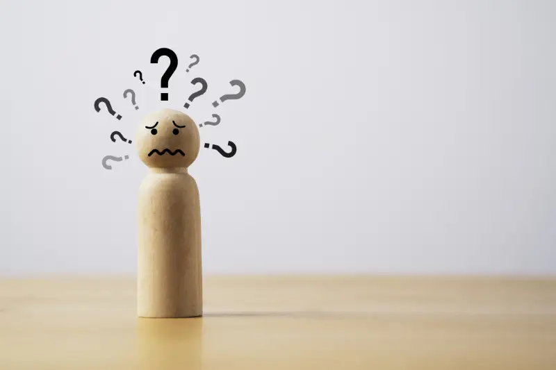 Holzfigur mit fragendem Gesichtsausdruck, umgeben von Fragezeichen, symbolisiert Missverständnisse in der Kommunikation auf heller Tischplatte vor grauem Hintergrund