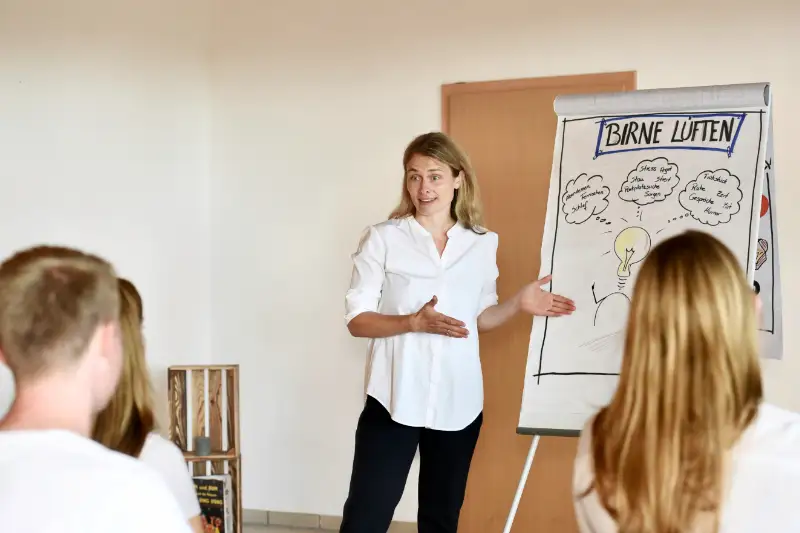 Coachin erklärt in einem Seminar zur Stressbewältigung ein Flipchart mit der Überschrift „Birne lüften“ vor mehreren Teilnehmenden