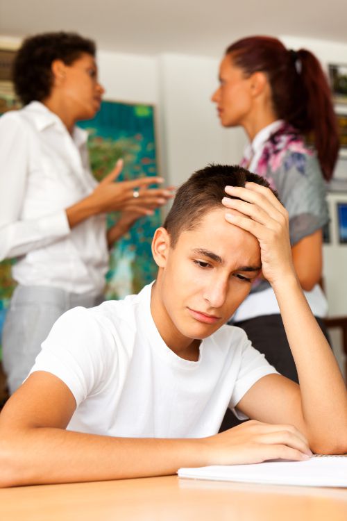 Jugendlicher sitzt frustriert am Tisch, im Hintergrund diskutieren zwei Erwachsene, symbolisiert Konflikte im Familienalltag vor einem Elterntraining in Bocholt